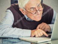 stock-photo-elderly-old-man-using-computer-sitting-at-table-isolated-on-grey-wall-background-senior-people-and-252858463