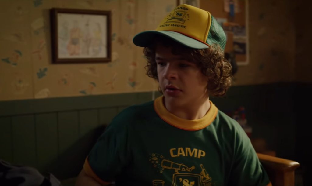 Dustin porte une casquette et un t Shirt « Camp Know Where » dans la saison 3. // Source : Youtube/Netflix Dustin porte une casquette et un t Shirt « Camp Know Where » dans la saison 3. // Source : Youtube/Netflix