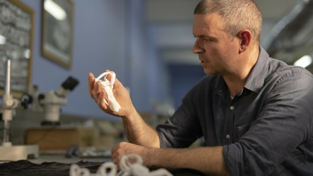 Le professeur Jonah Choiniere tenant entre ses mains un moulage 3D d'un os de Shuvuuia deserti, et exprimant sur son visage la surprise face à ce fossile incroyable. // Source : Wits University