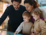 Un couple et ses enfants regardent une borne intéractive dans un centre commercial pour se repérer // Source : Midjourney - image générée par IA Un couple et ses enfants regardent une borne intéractive dans un centre commercial pour se repérer // Source : Midjourney - image générée par IA
